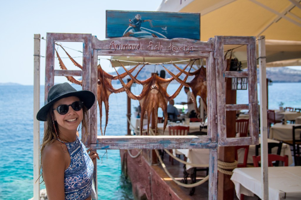 Bianca posing with an Octopus hanging on a frame in Ammoudi Bay, Santorini. Discover the unique experiences of Santorini with our travel blog.