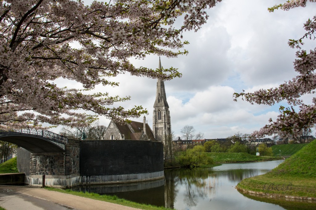 View of the Fortress in Copenhagen, Denmark - a serene park with lush greenery, a nearby church, and a beautiful fountain. Enjoy a peaceful retreat in the heart of the bustling city.