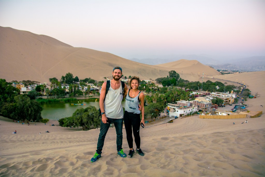 Image featuring Bianca and Brett posing in front of the picturesque village of Huacachina, showcasing its charm amidst desert landscapes in Peru.