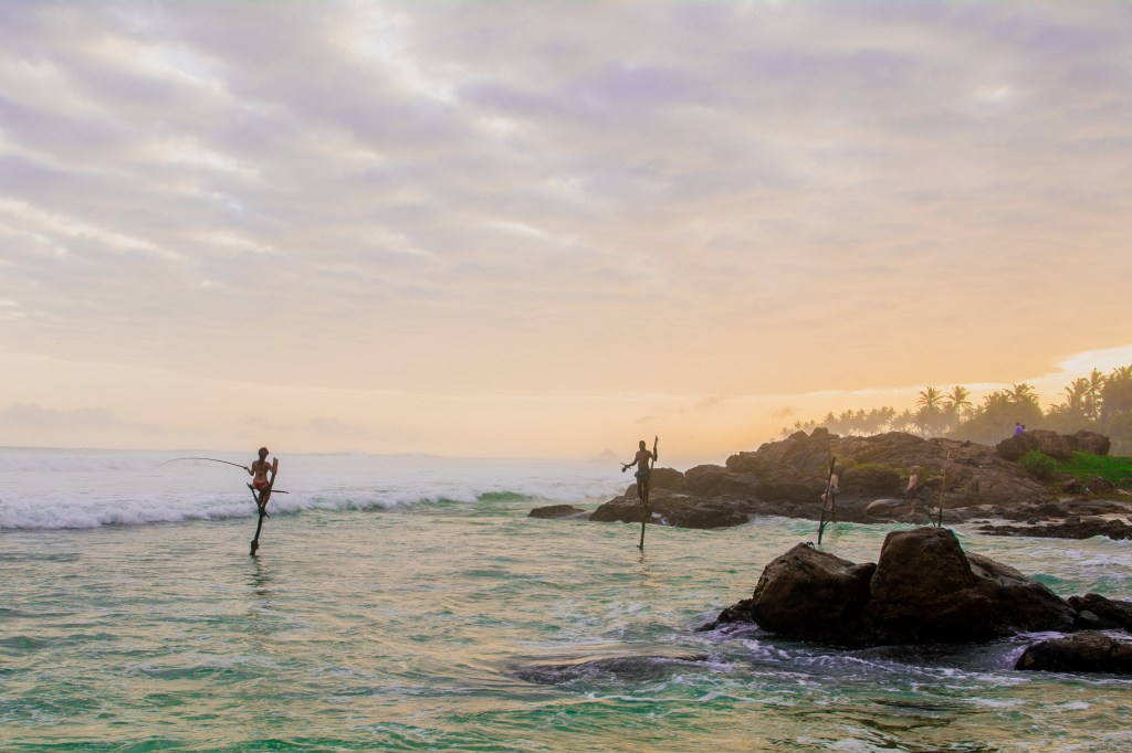 Image showcasing local fishermen perched on sticks over the coral, fishing for their families and market sales.