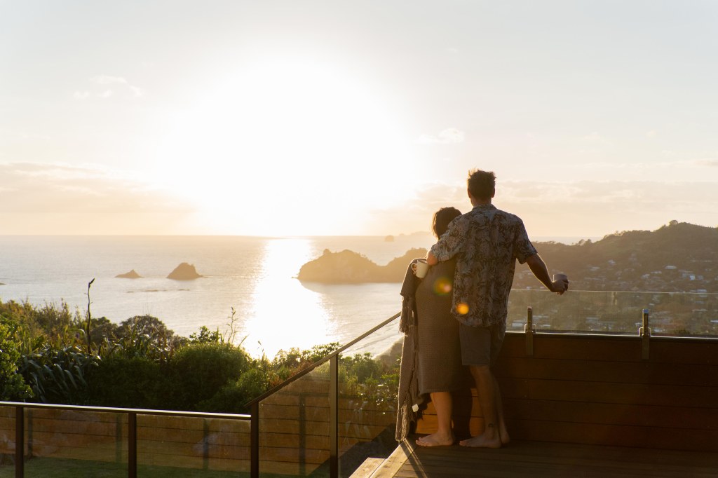 Bianca and Brett enjoying morning coffee at sunrise with a scenic view of Hahei Beach on their New Zealand travel adventure.
