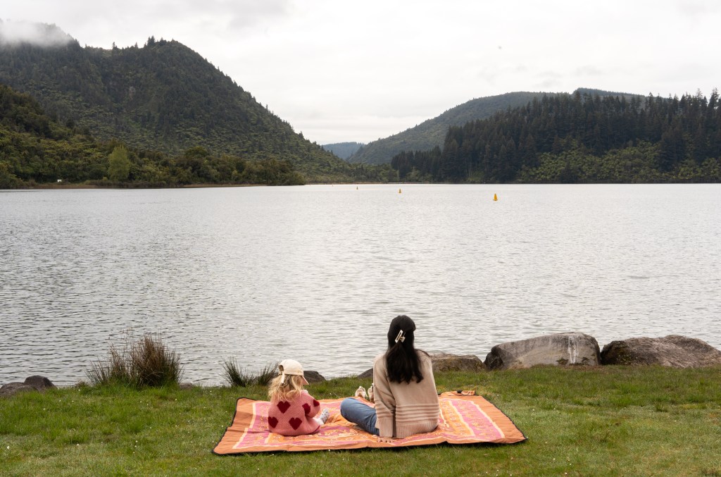 Bianca and Florence enjoying a coffee and fluffy while overlooking the serene Blue Lake (Lake Tikitapu) in Rotorua, New Zealand — peaceful family moment during a lakeside getaway.